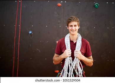 Active young man in sportswear standing with rope on shoulders against artificial training climbing wall. Smiling climber with cord, harness, carabiners and equipment looks to the camera outdoors.