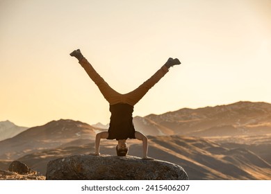 Active young man is standing upside down on big rock in sunset mountains