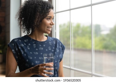 Active young mixed race lady stand by window at home hold glass of fresh pure water. Smiling black woman enjoy good habit drink mineral aqua at morning take care of skin body health beauty. Copy space