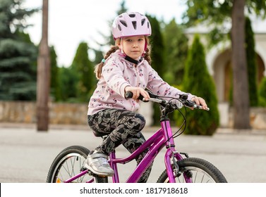 Active young school age girl, child riding a bike in a helmet, closeup. Kid rides a bicycle on a sunny day. Hands on the handle bars, looking at the camera, portrait shot. Fun outdoors activities