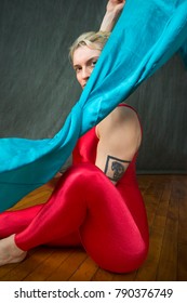 Active Young Woman Dancer With Short Blonde Hair, Seated On The Floor In A Red Unitard, Waving A Blue Scarf Overhead In The Studio.