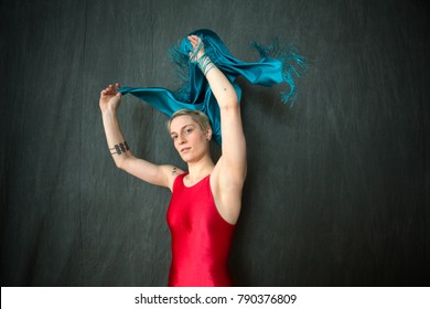 Active Young Woman Dancer With Short Blonde Hair, In A Red Unitard Waving A Blue Scarf Overhead In The Studio.