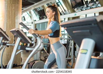 Active young woman exercising on elliptical trainer machine at gym. Sporty smiling girl listening to music and working out on elliptical trainer machine wearing sportswear in gym. - Powered by Shutterstock