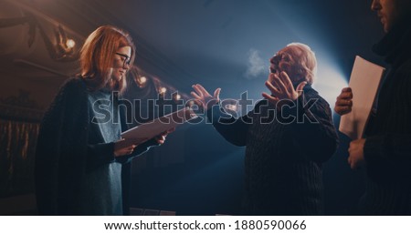 Actor and actress with scripts holding hands and speaking with each other while rehearsing romantic scene with director on stage in theater