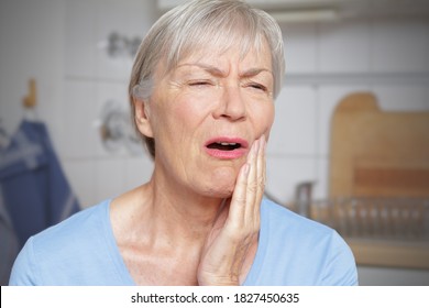 Acute toothache: elderly woman in her kitchen with a hand at her painful cheek.