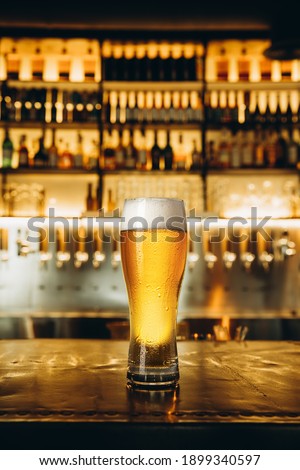 Ad. Full glass of light lager beer with drops on the side, wooden table in warm light of bar. Alcohol, entertainment, traditional drinks, Oktoberfest atmosphere concept. Modern bar, vertical crop.
