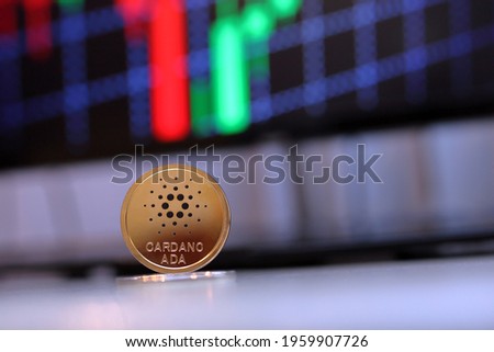 Ada cardano coin sitting on a desk with a red and green background chart computer pc screen