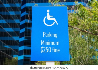 ADA Handicapped Sign With Handicapped Wheelchair Symbol) Marks Accessible Parking Space  On Outdoor Parking Lot. Minimum Fine $250 Warning Sign. Green Landscaping And Office Buildings In Background