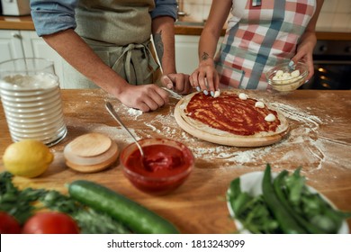 Add cheese. Cropped shot of couple making pizza together at home. Man in apron adding, applying tomato sauce on the dough while woman adding mozzarella. Selective focus. Horizontal shot