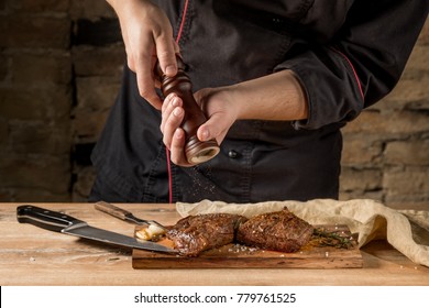 Add Pepper To Beef Steak With Mill In Chef Hands, Black Background. Food Recipe Photo, Copy Text.