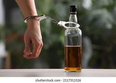 Addicted man in handcuffs with bottle of alcoholic drink at table against blurred background, closeup