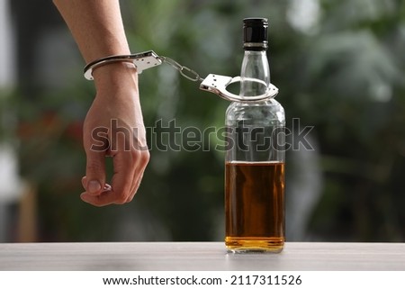 Addicted man in handcuffs with bottle of alcoholic drink at table against blurred background, closeup