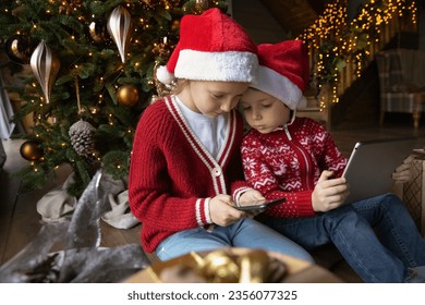 Addicted to modern technology happy little kids siblings using different gadgets, involved in playing online games, watching funny cartoons, spending time online sitting near decorated Christmas tree. - Powered by Shutterstock