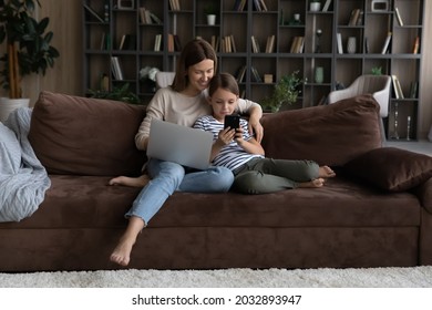 Addicted to modern technology happy young mother and adorable little kid daughter using different gadgets, playing entertaining spending time online resting on comfortable sofa in living room. - Powered by Shutterstock