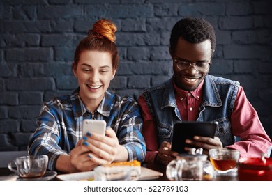 Addicted To Social Networking. Couple Using Modern Gadgets While Relaxing At Cafe. Red-haired Girl Reading Information On Web Page Via Mobile Phone While Black Man Watching Video On Digital Tablet