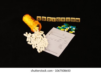 Addiction Spelled Out With Tiles Above Spilled White And Colored Pills Over A Prescription Pad On A Black Background.
