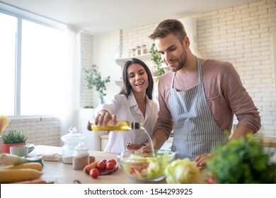 Adding Olive Oil. Beautiful Young Wife Adding Some Olive Oil To Salad While Cooking With Husband