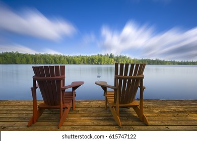 Adirondack Chairs On A Lake Deck
Two Adirondack Chairs Sitting On A Wood Dock Facing A Lake. The Wood On The Dock Is Arranged In An Horizontal Pattern. On One Of The Chairs There’s A Glass Of Wine.