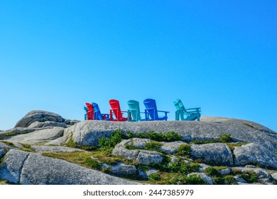 Adirondack chairs, two blue, two green, and two red on the edge of a rocky cliff overlooking the ocean on a sunny summer's day. The sky is blue and clear. The rocky cliff has cracks and spaces. - Powered by Shutterstock