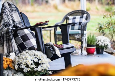 Adirondack rocking chair with style buffalo check blanket and pillows on a porch or patio decorated for autumn with heirloom gourds & white and orange mums. Selective focus with garden in background.