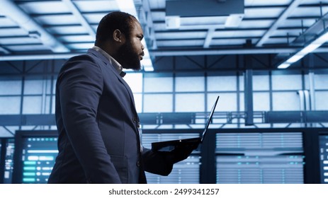 Admin doing yearly checkup using notebook to prolong data center electronics life span and avoid downtime. Man walks in server room, monitoring server infrastructure components using laptop, camera A