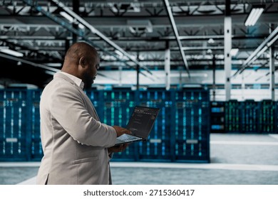 Admin walking in data center running lines of code on laptop, troubleshooting rackmounts. African american IT expert using programming scripts on notebook device in server room
