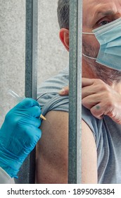 Administration of COVID-19 vaccine in a prison. A middle-aged man in a mask receives intramuscular injection through bars of his cell.