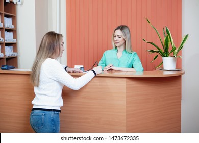 Administrator And Patient At The Reception In The Medical Office. Young Female Receptionist With Client In Hospital.