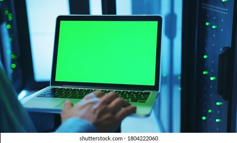 IT administrator using a mock-up laptop running diagnostics and web hosting with chrome key greenscreen device inside server cabinet of database. Close-up. Advanced data center.