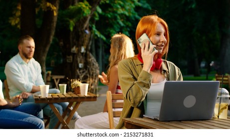 An Admirable Woman With Long Ginger Hair Is At A Table Near A Cafe In The Park, She Is In Deep Conversation On Her Phone Looking Very Happy While Doing Hand Motions, In The Back A Group Of Friends Are