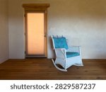An adobe style interior with corner in view, likely a bedroom or sitting room. Glass door to the outside, wide plank hardwood floor. Antique white wicker rocker completes the look. Santa Fe NM 2026