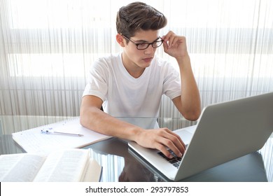 Adolescent young man at a glass desk at home doing his homework using a laptop computer, wearing spectacles and being concentrated, interior. Teenager boy studying against large window, indoors.