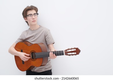 Adolescents, hobbies and pastime. A modern teenage boy in glasses and a grey T-shirt plays an acoustic guitar on a white background with copy space. 