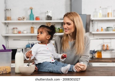 Adopted African American Kid Holding Spoon Near Bowl While Sitting On Kitchen Table Near Happy Mother