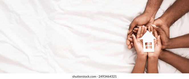 Adoption and family housing concept. Cutout paper house in hands of african american family over white bed sheet background. Closeup, panorama - Powered by Shutterstock