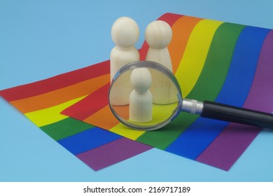 Adoption For LGBT Family Concept. Wooden People Figures Of Father, Mother And Child With Magnifying Glass On LGBT Flag.