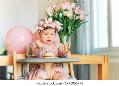 Adorable 1 year old baby girl eating cupcake, happy child sitting in a chair, tasting sweet dessert, first birthday
