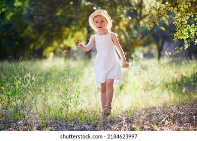 Adorable 4 Year Old Girl In White Dress And Straw Hat Walking In A Garden In Provence, France