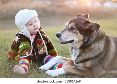 An adorable 8 month old baby girl is bundled up in a sweater and bomber hat looking lovingly at her pet German Shepherd dog as they sit outside on a cold fall day.