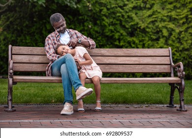 Adorable African American Girl And Her Grandfather Sitting On Bench In Park  