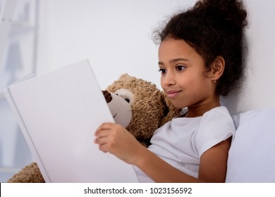 Adorable African American Kid Reading Book And Hugging Teddy Bear At Home