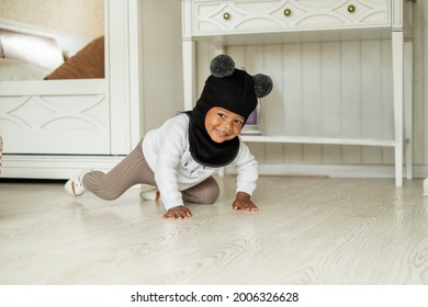 Adorable african american little boy in hat helmet with two fluffy bobbles crawling on floor of bedroom and laughing, having fun trying new accessoire, child fashion