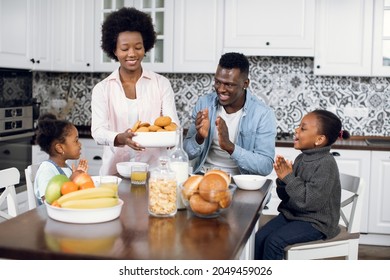 Adorable african american women serving on table tasty homemade cookies for her lovely family. Two happy daughters and handsome husband sitting together on modern kitchen and clapping hands