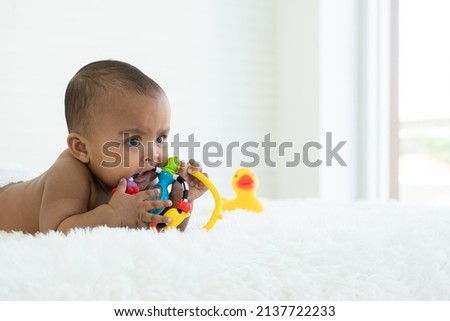 Adorable African Asian newborn girl lying on bed holding and biting toy at home. Baby teething concept. Copy space