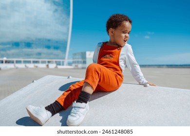 Adorable african baby child in orange overalls, sits in a ramp looking away, outdoors by the sea at city. Happy girl playing alone. Copy space. - Powered by Shutterstock