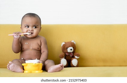Adorable African baby newborn in diaper sitting on sofa with small bear doll trying to grab a spoon to feed herself. Spilled food messy in mouth and body of little kid. Child development concept - Shutterstock ID 2152975403