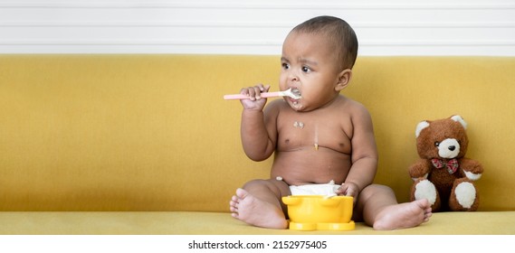 Adorable African Baby Newborn In Diaper Sitting On Sofa With Small Bear Doll Trying To Grab A Spoon To Feed Herself. Spilled Food Messy In Mouth And Body Of Little Kid. Child Development Concept