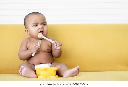 Adorable African Baby Newborn In Diaper Sitting On Yellow Sofa Trying To Grab Two Spoons To Feed Herself. Spilled Food Messy In Mouth And Body Of Little Kid. Child Development Concept