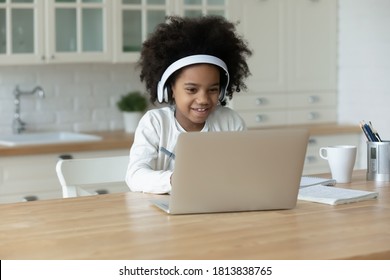 Adorable african ethnicity small girl wearing headphones, looking at computer screen. Happy cute mixed race child enjoying online educational lecture or learning using laptop software application.