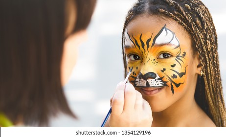 Adorable african-american girl getting tiger face painting in park, empty space